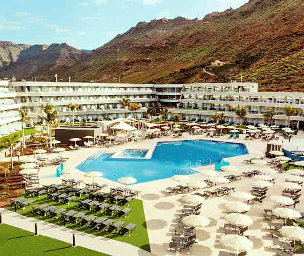 Et hotelområde med en stor udendørs swimmingpool, liggestole og parasoller, omgivet af bygninger og bjerge i baggrunden. (AI-genereret tekst)