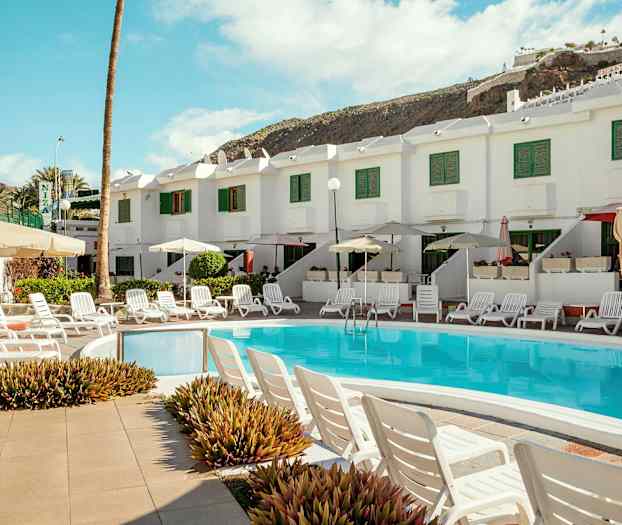 Poolområde med liggestole og parasoller foran et hvidt hus med grønne skodder. (AI-genereret tekst)