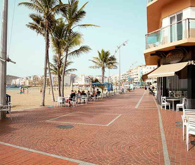 En strandpromenade med palmer, cafestole og bygninger i baggrunden. (AI-genereret tekst)