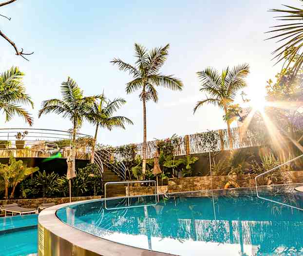 En klar pool med palmer og udendørs servering i baggrunden under en solrig himmel. (AI-genereret tekst)