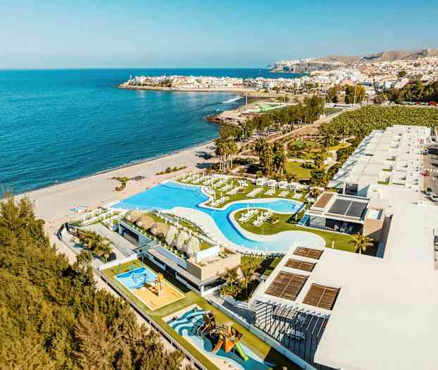 Luftfoto af en kystanlæg med poolområde og bygninger ved siden af en sandstrand og havet. (AI-genereret tekst)