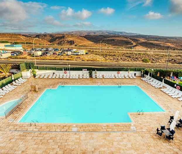 En stor udendørs pool omgivet af liggestole og parasoller. I baggrunden ses en vej og et landskab med bakker. (AI-genereret tekst)