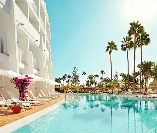 Hotellets facade med altaner, liggestole ved poolen og palmer i omgivelserne. (AI-genereret tekst)