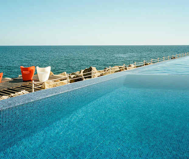 En infinitypool med udsigt over et roligt hav og et trægulv med farverige stole. (AI-genereret tekst)