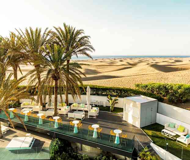 En terrasse med siddepladser og parasoller foran en ørken med sandklitter og palmer. (AI-genereret tekst)