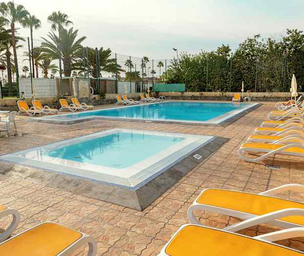 Udendørsområde med liggestole omkring to pools, omgivet af palmer og hegn. (AI-genereret tekst)