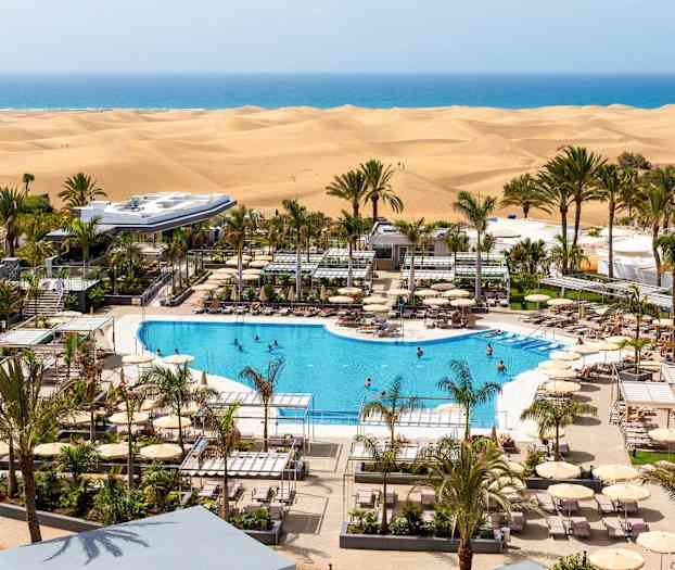 Udsigt over hotellets pool omgivet af liggestole og parasoller med sandklitter og hav i baggrunden. (AI-genereret tekst)