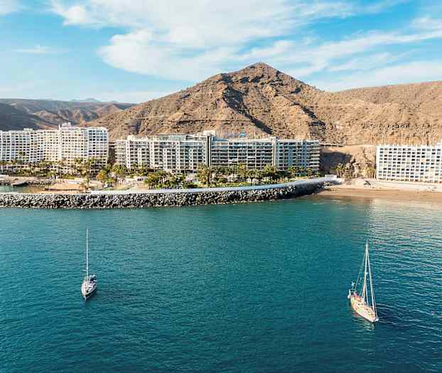 Udsigt over flere hotelbygninger ved havet med en bjergskråning i baggrunden og sejlbåde i vandet. (AI-genereret tekst)