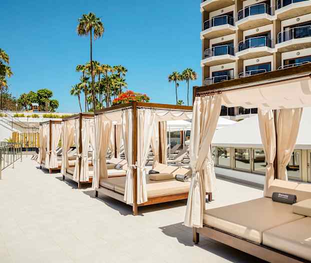 Billede af solsenge med gardiner ved en hotelpool, med palmer og en bygning i baggrunden. (AI-genereret tekst)