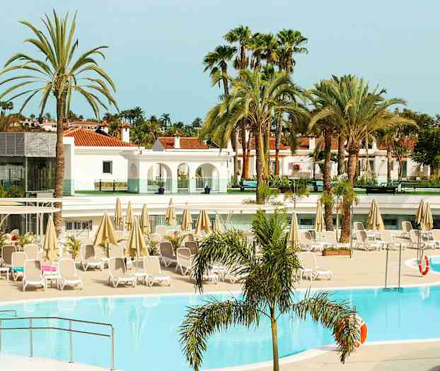 Poolområde ved et hotel med liggestole og parasoller, omgivet af palmer og bygninger. (AI-genereret tekst)