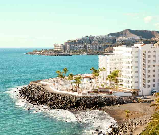 En hvid bygning ved en stenet kyst med palmer og hav i baggrunden. (AI-genereret tekst)