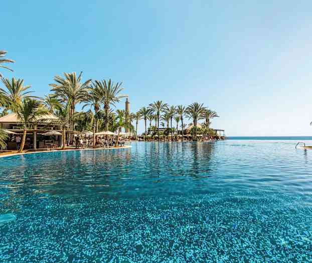 En stor pool omgivet af palmer og en blå himmel med havet i baggrunden. (AI-genereret tekst)