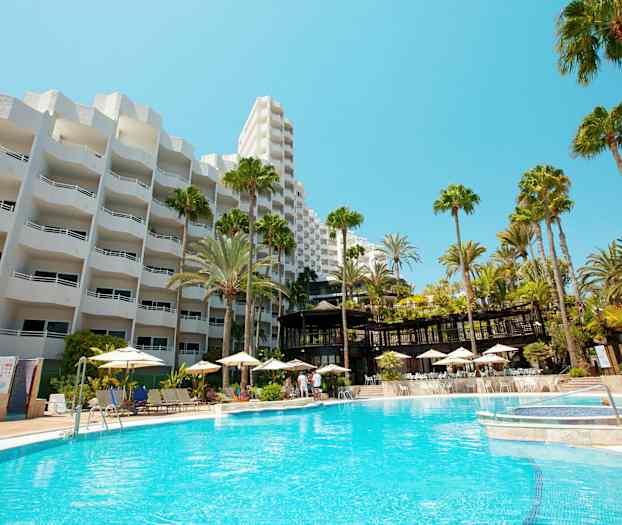 Hotelbygning med hvid facade og en stor udendørs pool omgivet af liggestole og parasoller. (AI-genereret tekst)