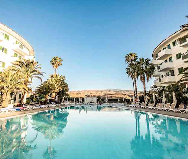 Et hotelområde med pool i forgrunden, omgivet af liggestole og parasoller samt omgivende bygninger på siderne. (AI-genereret tekst)