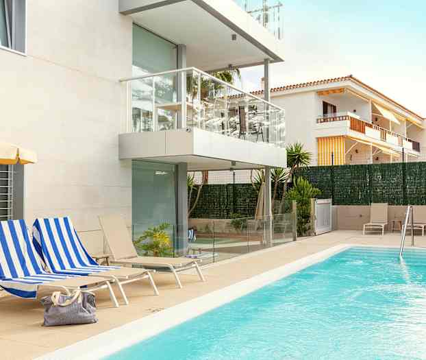 En udendørs swimmingpool ved et moderne lejlighedskompleks med hvid facade og altaner. Stole og liggestole er placeret rundt om poolområdet, og en taske ligger på en af liggestolene. (AI-genereret tekst)