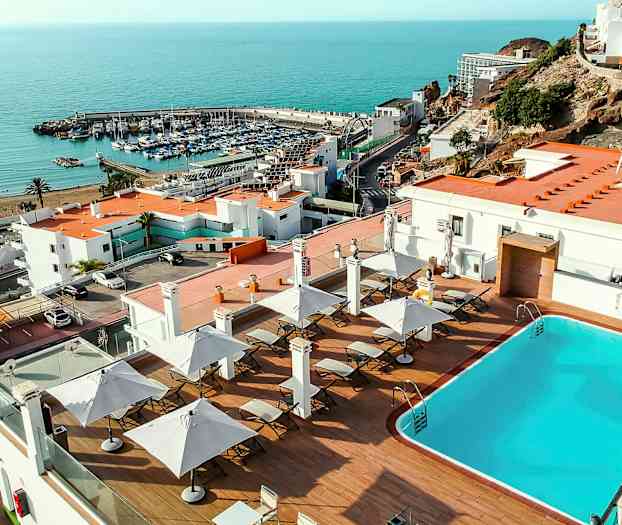 En pool med liggestole og parasoller på en terrasse med udsigt over havet og en marina. (AI-genereret tekst)