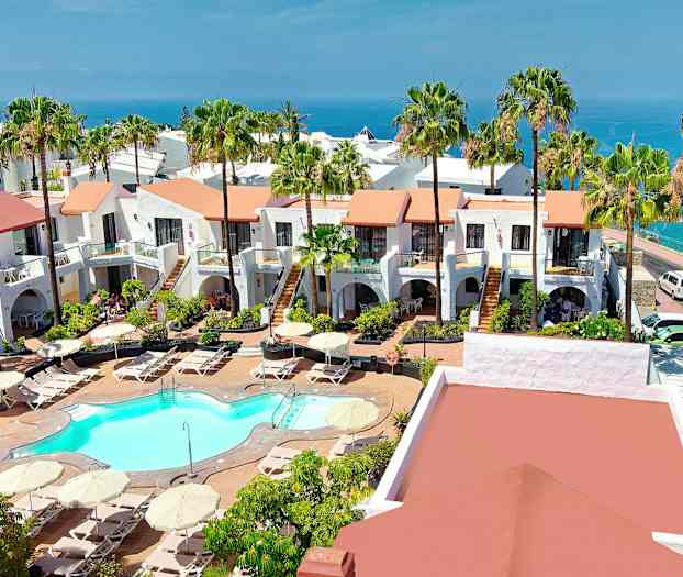 Udsigt over et hotelområde med hvidkalkede bygninger, en pool og frodige haver, havet kan ses i baggrunden. (AI-genereret tekst)