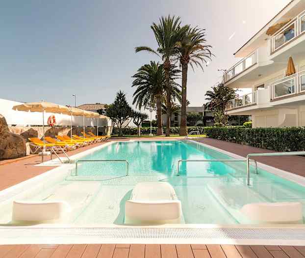 Poolområde med liggestole, parasoller, en stenmur og omkringliggende træer. (AI-genereret tekst)