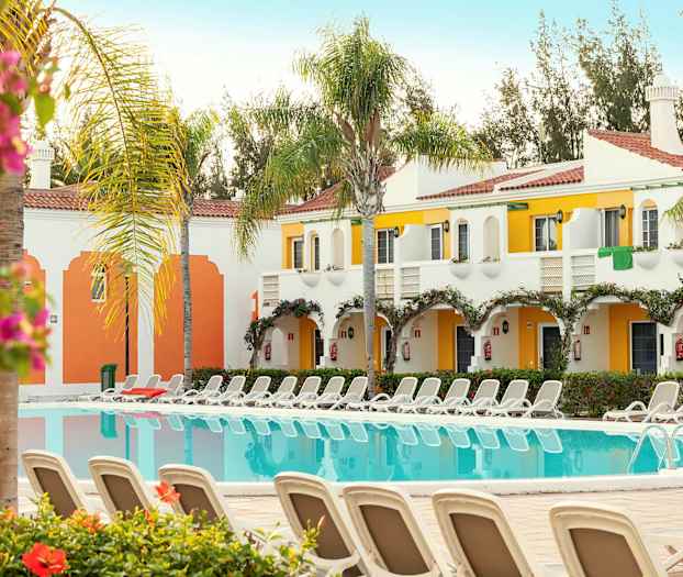 Et billede af en pool med liggestole omkring, omgivet af hvidmalede og farverige bygninger med altaner. Palme- og blomsterplanter ses også. (AI-genereret tekst)