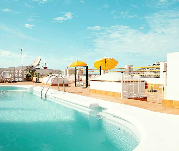 En udendørs pool med turkis vand omgivet af en solterrasse med gule parasoller. (AI-genereret tekst)