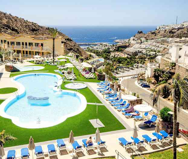 Poolområde med liggestole og parasoller, omgivet af bygninger og bjerge, med havet i baggrunden. (AI-genereret tekst)