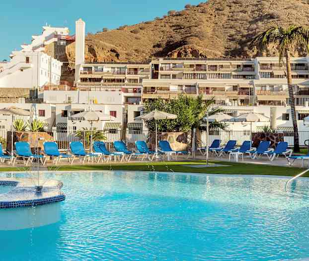 En pool omgivet af liggestole og parasoller foran en bygning på en skråning. (AI-genereret tekst)