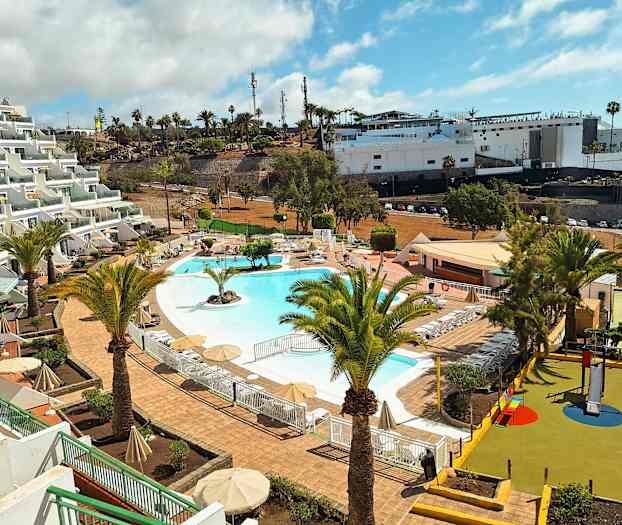 Billede af et feriested med en pool omgivet af liggestole og parasoller. I forgrunden ses palmer og en legeplads for børn. (AI-genereret tekst)