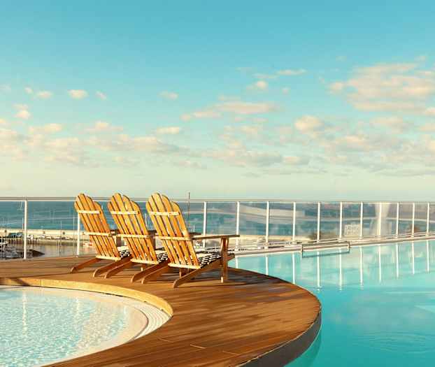 Tre træstole ved kanten af en pool med udsigt over havet. (AI-genereret tekst)