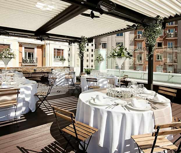 Billedet viser et elegant udendørsområde med opdækkede borde og træstole under en overdækket pergola. (AI-genereret tekst)