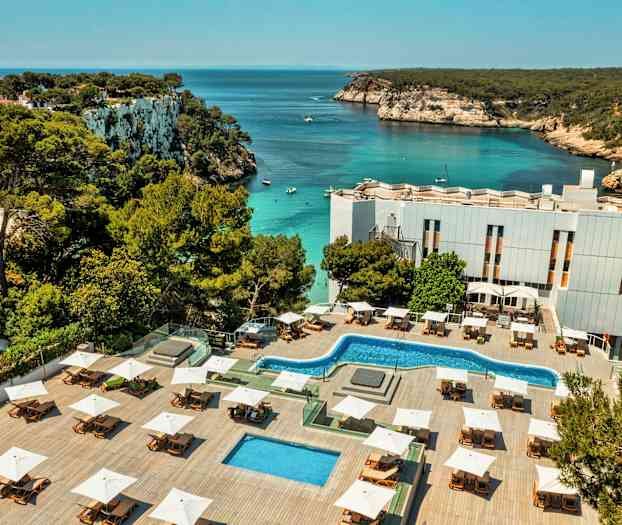 Et hotelområde med pools og liggestole ligger ved en bugt med klart vand og klippekyst. (AI-genereret tekst)