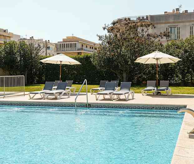 En udendørs swimmingpool med blåt vand har liggestole og parasoller på et flisebelagt område ved siden af græs og grønne buske. (AI-genereret tekst)