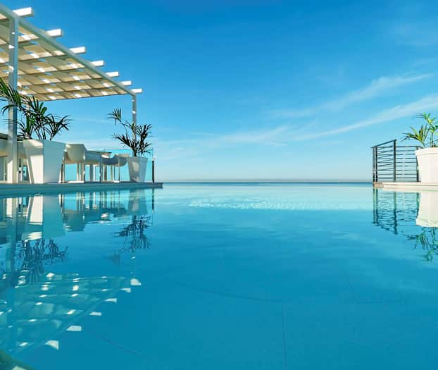 En infinity-pool med udsigt over havet, med borde og stole under en skyggekonstruktion ved siden af poolen. (AI-genereret tekst)