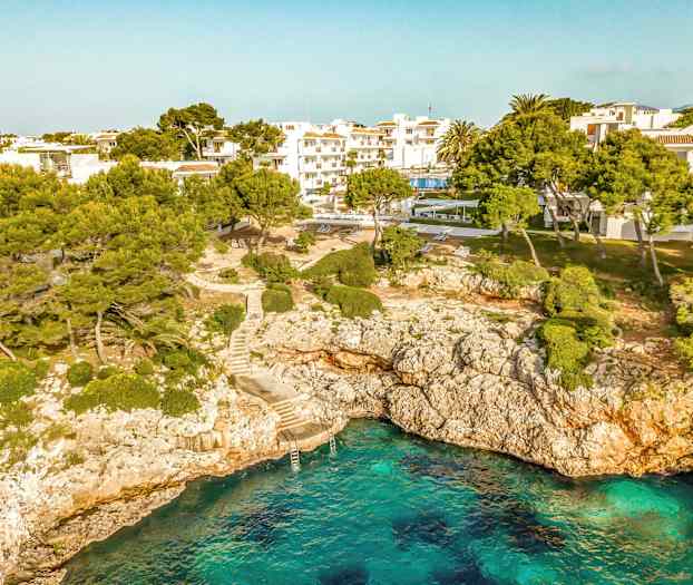 En kystlinje med klart blåt vand, klipper samt træer og bygninger i baggrunden. (AI-genereret tekst)