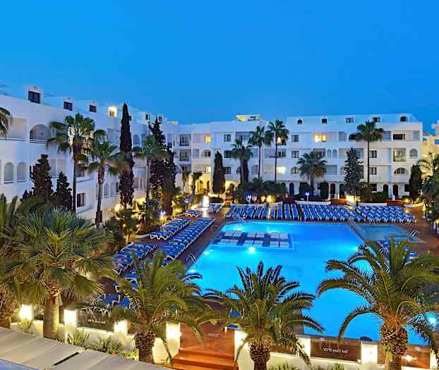 Et hotelområde om aftenen med en oplyst pool, omgivet af stole og grønne palmer. (AI-genereret tekst)