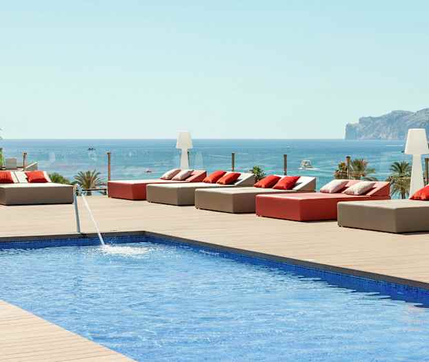 Udsigt over en pool med solsenge og puder på en træterrasse, med havet og et bjerg i baggrunden. (AI-genereret tekst)