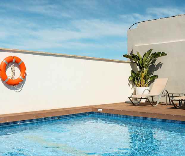 En udendørs pool med to liggestole og en orange redningskrans hængende på en hvid væg. (AI-genereret tekst)