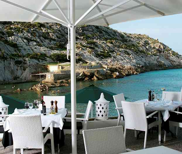 En udendørs restaurant med dækkede borde og hvide stole med udsigt til et klippefyldt kystlandskab og havet. (AI-genereret tekst)