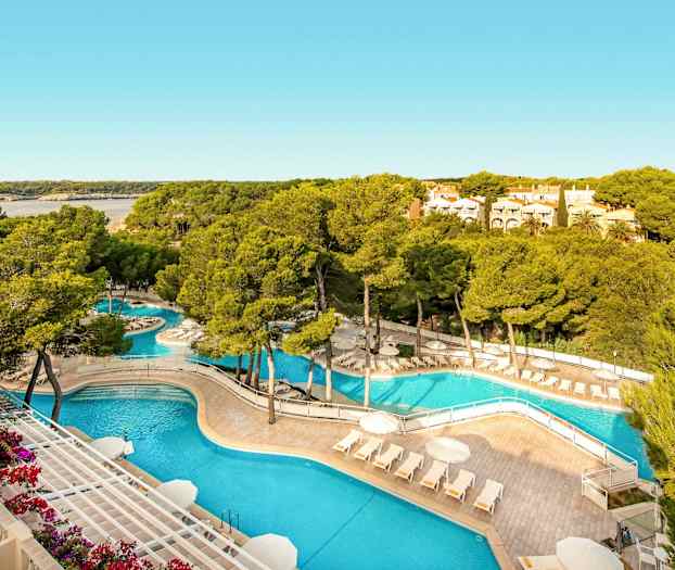 Et poolområde med liggestole og parasoller omgivet af træer og grønne områder, med en bugt og boliger i baggrunden. (AI-genereret tekst)