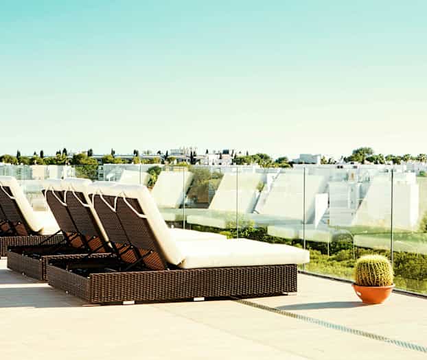 En terrasse med liggestole og glaskrækværk samt udsigt over en moderne bebyggelse og grønne områder. (AI-genereret tekst)