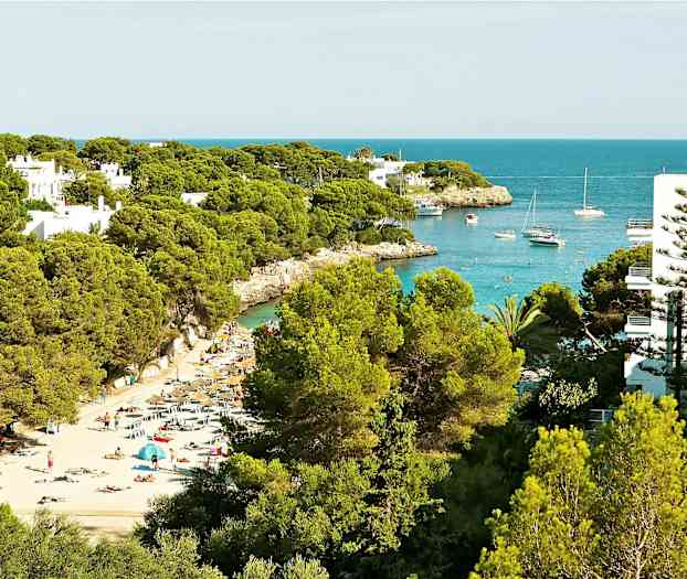En strand med liggestole og parasoller blandt frodige træer, og en bugt med både og klart blåt vand. (AI-genereret tekst)