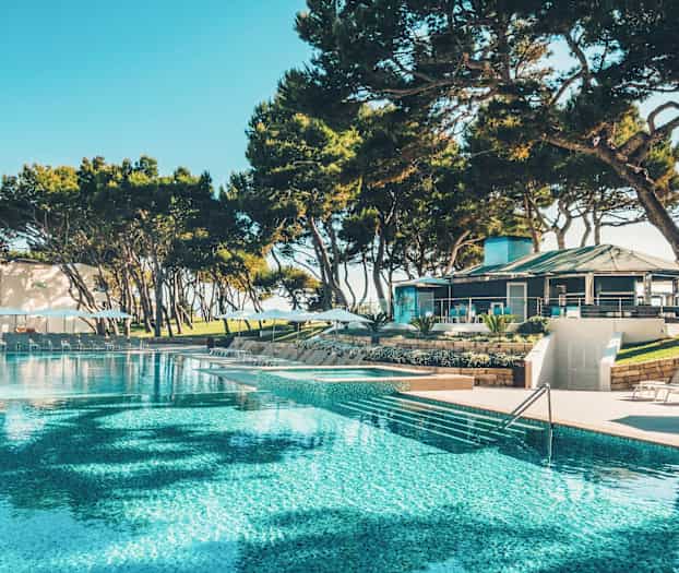 En stor pool omgivet af liggestole og parasoller, med en bygning og træer i baggrunden. (AI-genereret tekst)