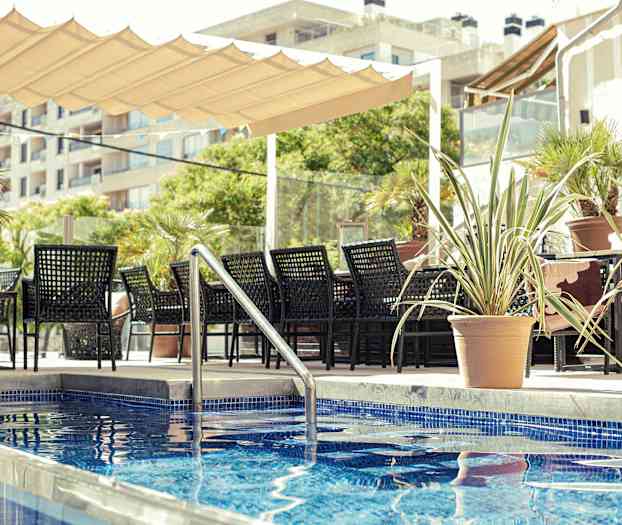 En siddeområde med sorte møbler ved siden af en pool med blå fliser, omgivet af potteplanter. (AI-genereret tekst)