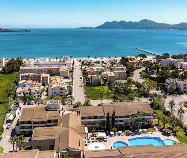 Luftfoto af en kystby med hotel, boliger og pool i forgrunden, med blåt hav og bjerge i baggrunden. (AI-genereret tekst)