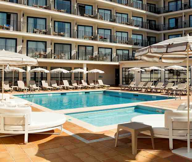 Et hotelpoolområde med liggestole, parasoller og en pool omgivet af flere etager med altaner. (AI-genereret tekst)