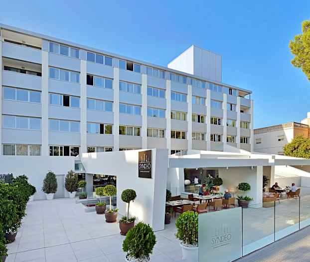 Billede af en moderne hotelbygning i flere etager med en udendørs servering og planter foran. (AI-genereret tekst)