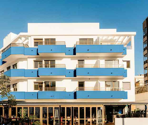 En lejlighedsbygning med hvide vægge og blå altaner, samt en restaurant i stueetagen. (AI-genereret tekst)