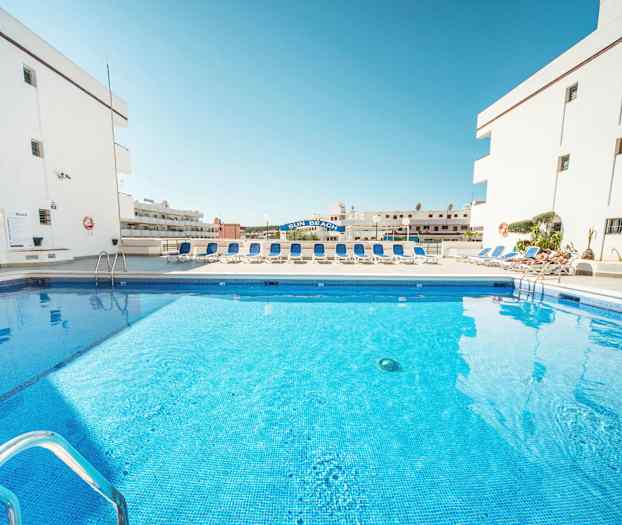 Billede af en swimmingpool omgivet af liggestole og hvide bygninger. (AI-genereret tekst)