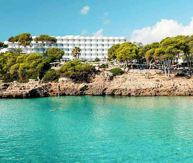 Hotel med hvide bygninger bag en klippefyldt strand med klart blåt vand. (AI-genereret tekst)