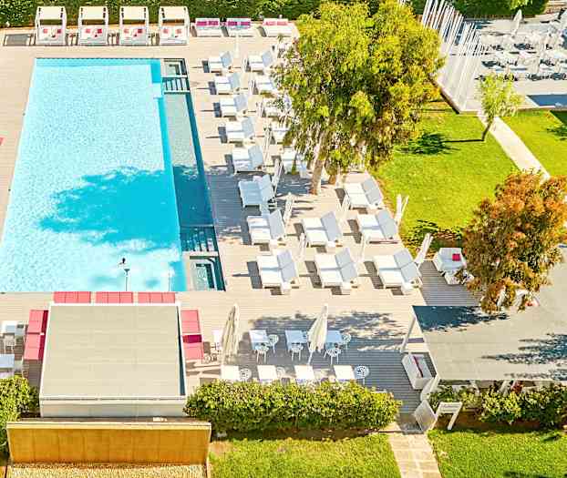 Udsigt oppefra over en udendørs swimmingpool omgivet af liggestole og grønne områder. (AI-genereret tekst)