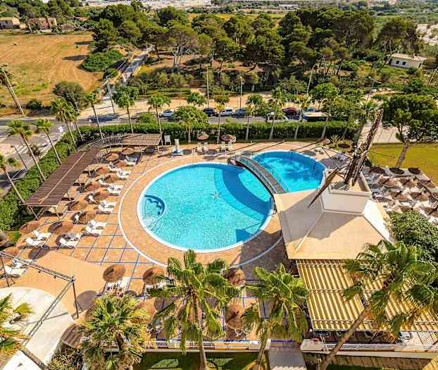 Luftfoto af et udendørsområde med to swimmingpools, liggestole og parasoller, omgivet af grønne arealer. (AI-genereret tekst)